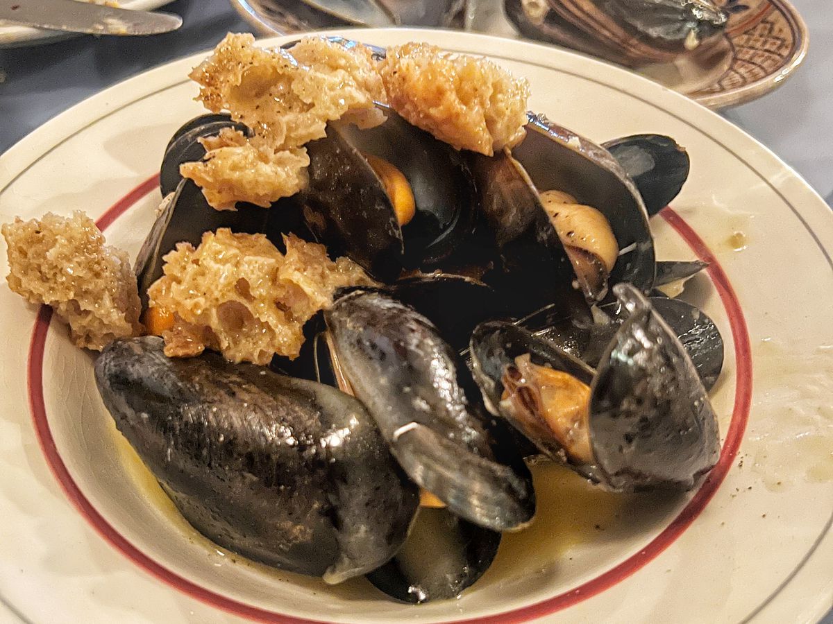 A white plate piled high with cooked mussels, the orange flesh nestled in the blue shells, with chunks of bread soaked in butter, garlic, and the cooking juices.