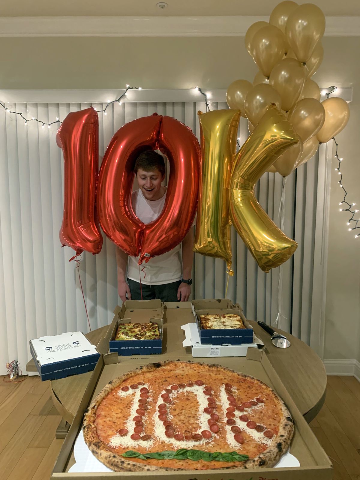 George Matelich stands with balloons that spell out “10K” alongside a pizza with “10K” spelled out in pepperoni, in honor of reaching 10,000 followers.