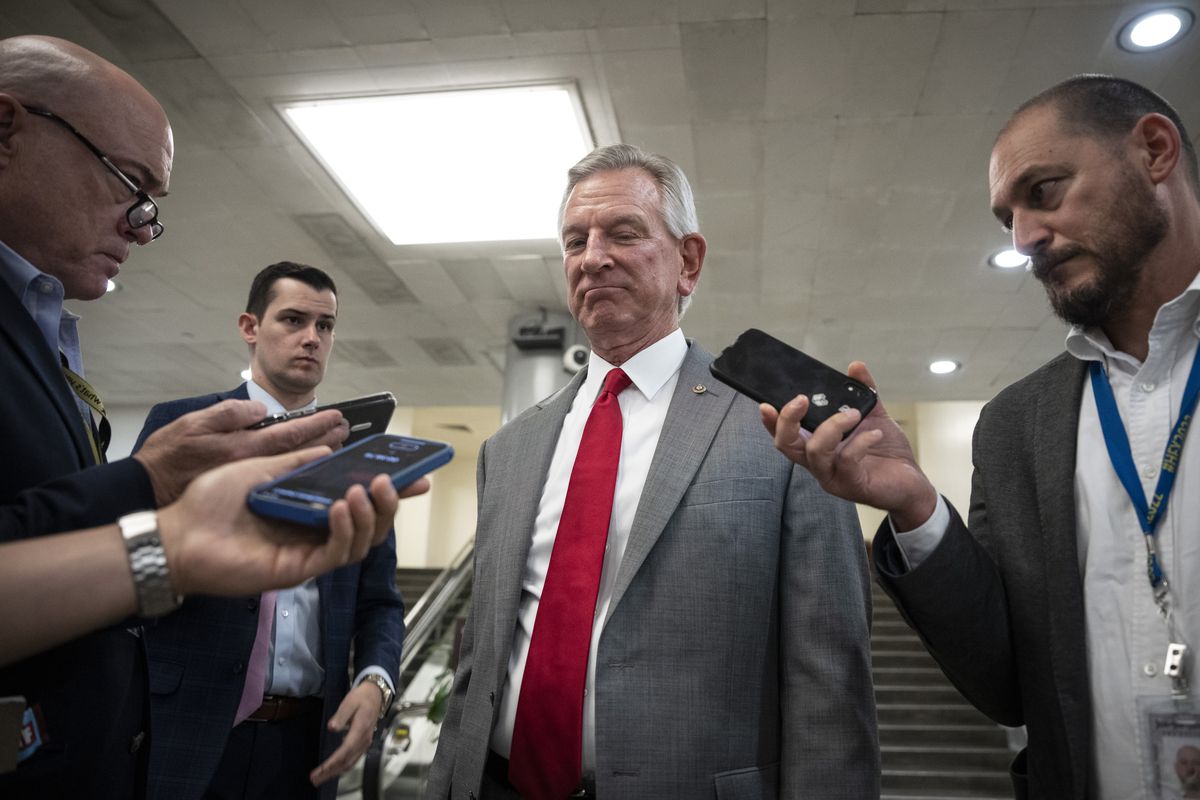 Senator Tommy Tuberville stands in the Senate subway in Washington, DC, wearing a grey suit and red tie. He is surrounded by several reporters holding out phones. 