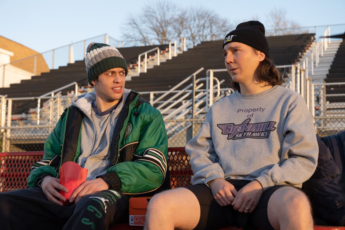 brothers Keith and Kevin Gill (Paul Dano and Pete Davidson), in slouchy casual wear (sweatshirts, hoodies, knit hats) sit on outdoor bleachers together in Dumb Money