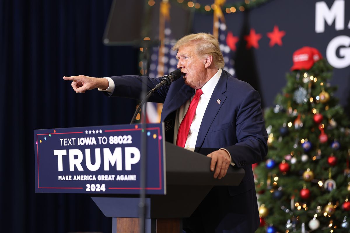 Trump stands at a podium and speaks energetically into a microphone, pointing a finger at the crowd. Behind him, a decorated Christmas tree is topped with a red baseball cap.