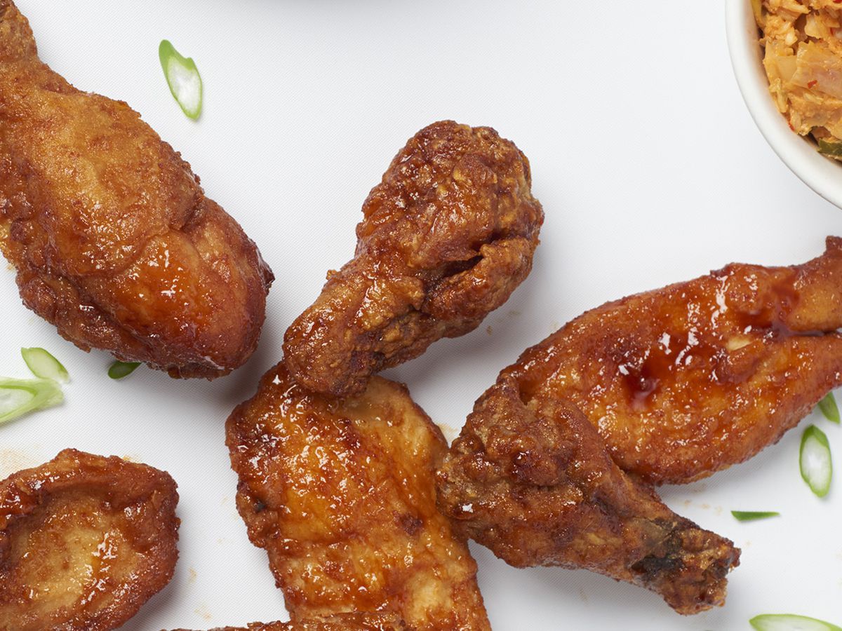 A spread of hot chicken wings and drumsticks on a white background, with a small bowl of kimchi visible in the corner of the photo