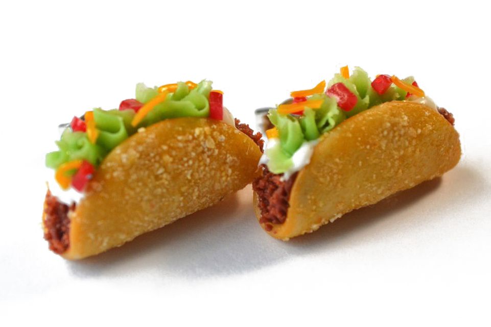 Two tiny clay tacos topped with sour cream and lettuce, with earring posts slightly visible coming out of the back, are on a white background 