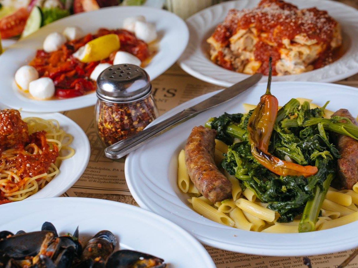 On a wooden table, several white dishes are partially visible, most holding different types of pasta including lasagna, penne with greens, and spaghetti with meatballs and red sauce.
