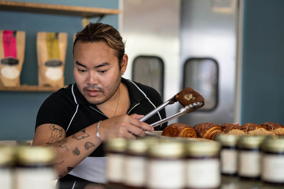 A man with a ponytail and arm tattoos pulls a pastry with metal tongs inside a sunny daytime bakery in Los Angeles.