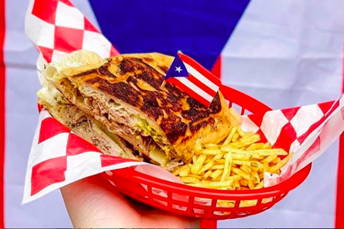 A hand holding a red basket containing a Mr. Pig sandwich with slow-roasted pernil, Swiss cheese, mustard, mayo, and pickles on a pan sobao