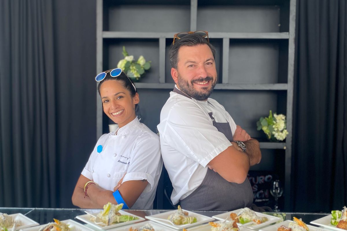 Chefs Alexandra Peña and Aaron Bludorn stand back-to-back with arms crossed in front of various plated dishes.