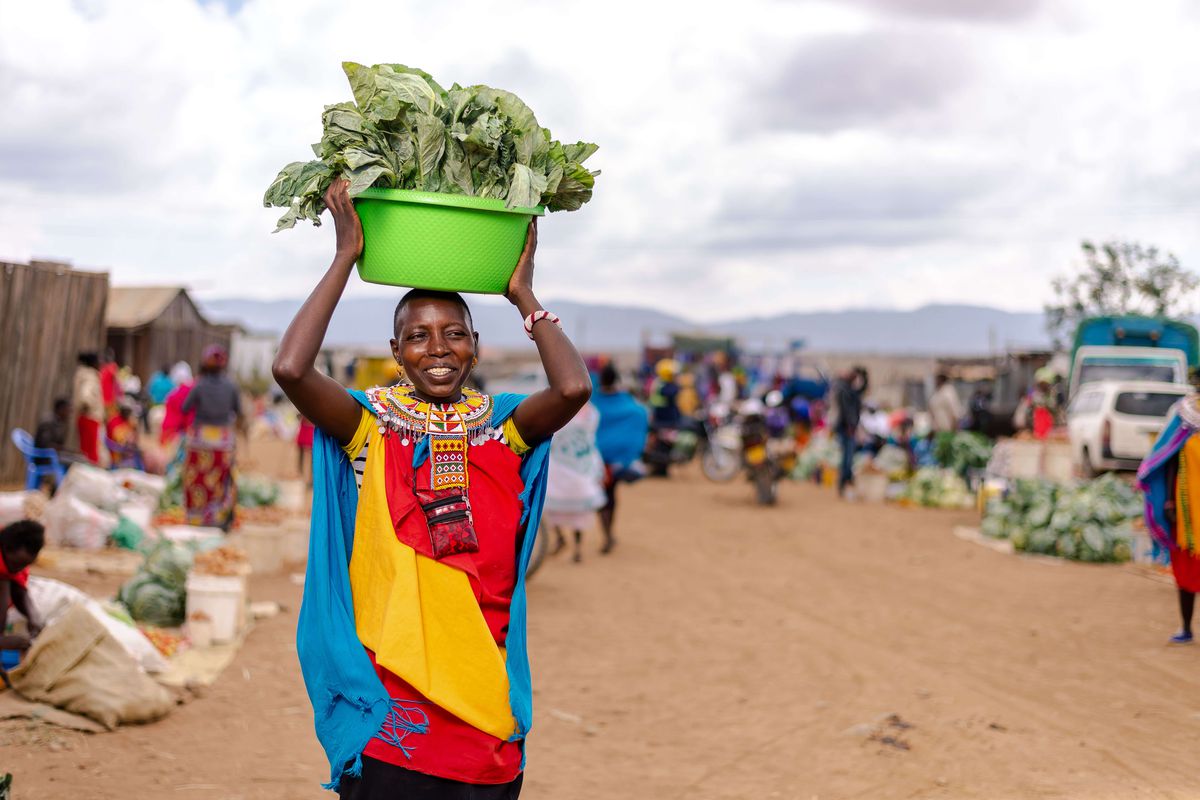 A woman in a colorful dress holds a bucket of greens on her head at an outdoor market in Kenya.