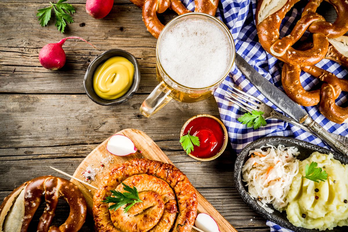 A top-down view of a food and drinks spread, with pretzels, a stein filled with beer, sausages, coleslaw, and potato salad