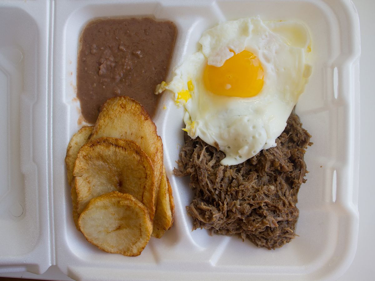 A container of barbacoa meat, sunny-side up fried egg, sliced potatoes, and refried beans. 