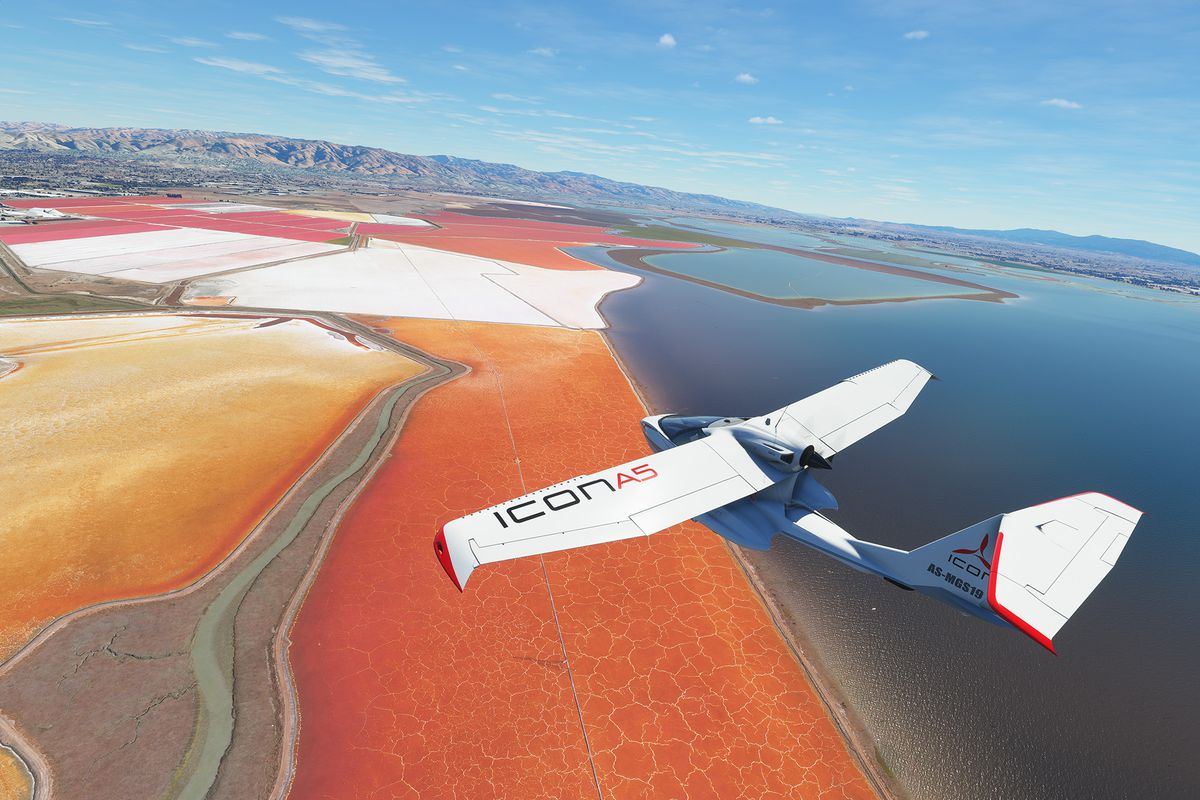 A white Icon A5, a two-seater aircraft with folding wings and a push-propeller configuration, flies over an arid salt pan on the edge of the ocean. The ground is brightly colored red and the sky is clear and blue.