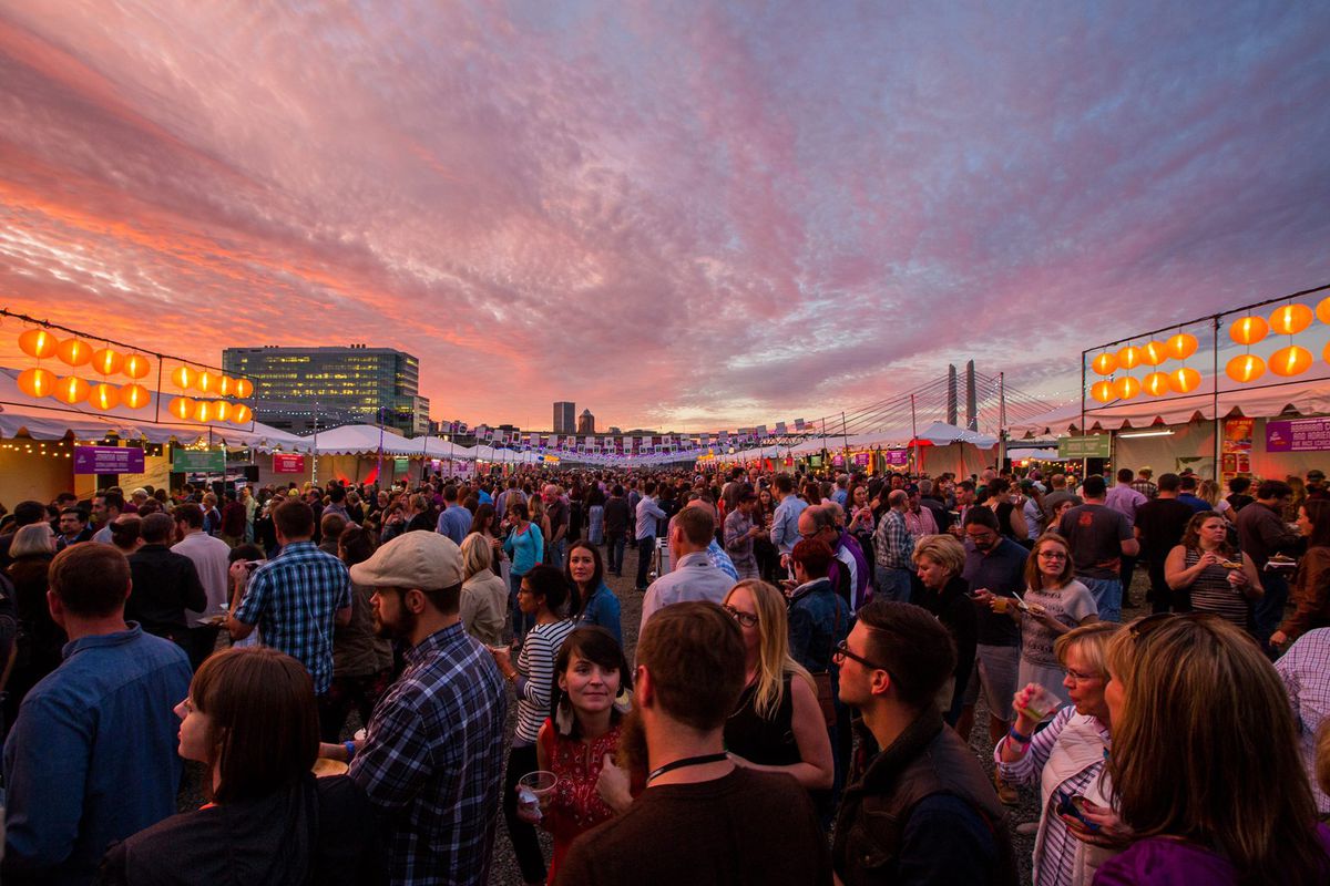 A massive crowd  of festival-goers stand in Feast’s Night Market, with lines of paper lanterns above a line of stalls. Sunset falls over the Tilikum Bridge in the background
