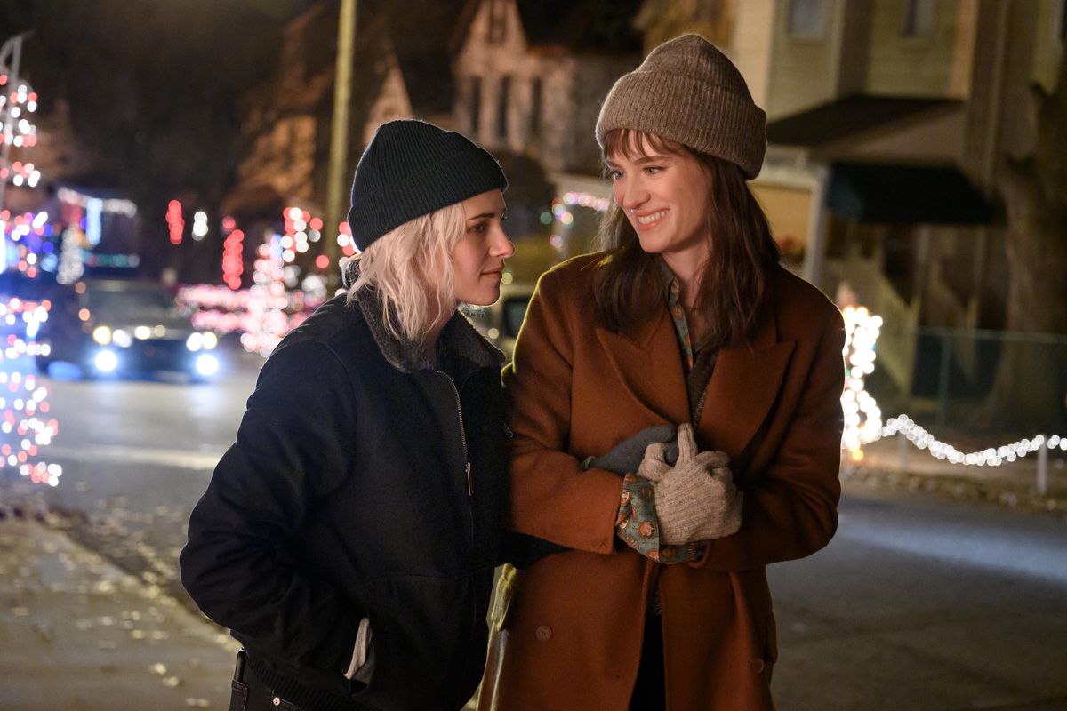 Two young women walk down a city street at Christmastime holding hands, dressed in winter clothing.