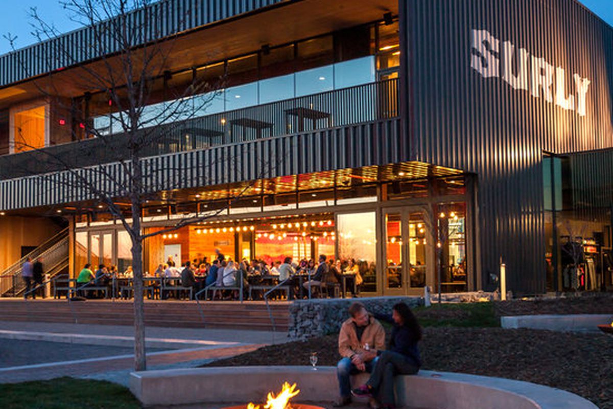 An exterior late summer shot of Surly’s monolith brewery. The bottom floor is filled with people inside. One couple is outside, near a fire pit by its giant outdoor green space.
