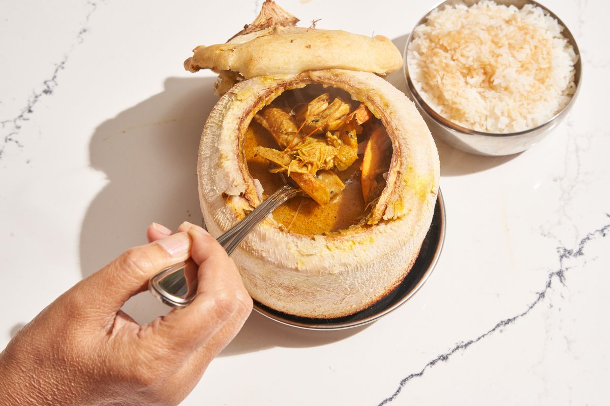 A hand sticks a spoon inside a beige coconut shell with the liner removed to reveal the food and sauce inside.  A cup of white rice is on the side.
