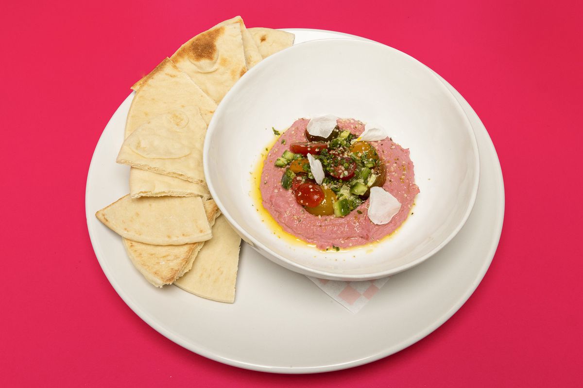 A bowl of pink dip with vegetables beside a plate of pita triangles.