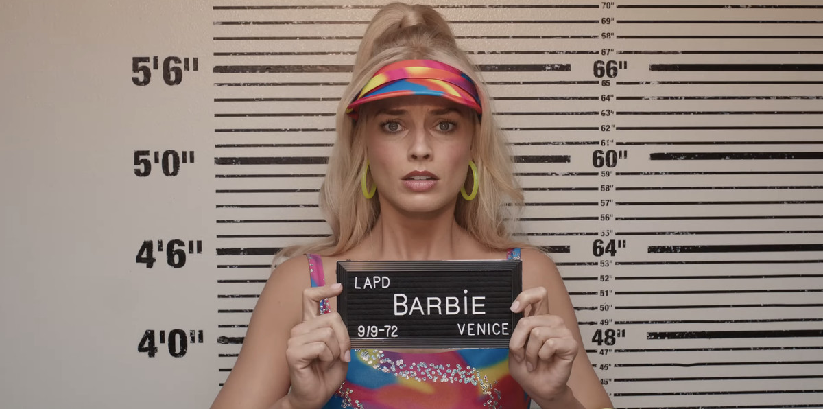 A still from the Barbie movie that shows Barbie taking a mugshot. Barbie is played by Margot Robbie and she looks distressed as she holds up a sign that says Barbie LAPD Venice.