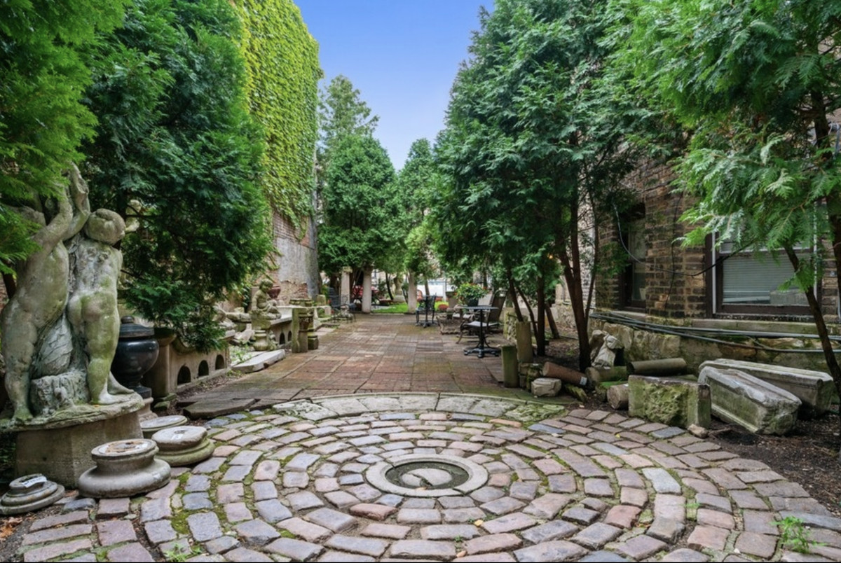 A side yard with lush trees, a vine-covered wall, a mossy statue, and a circular cobblestone path.