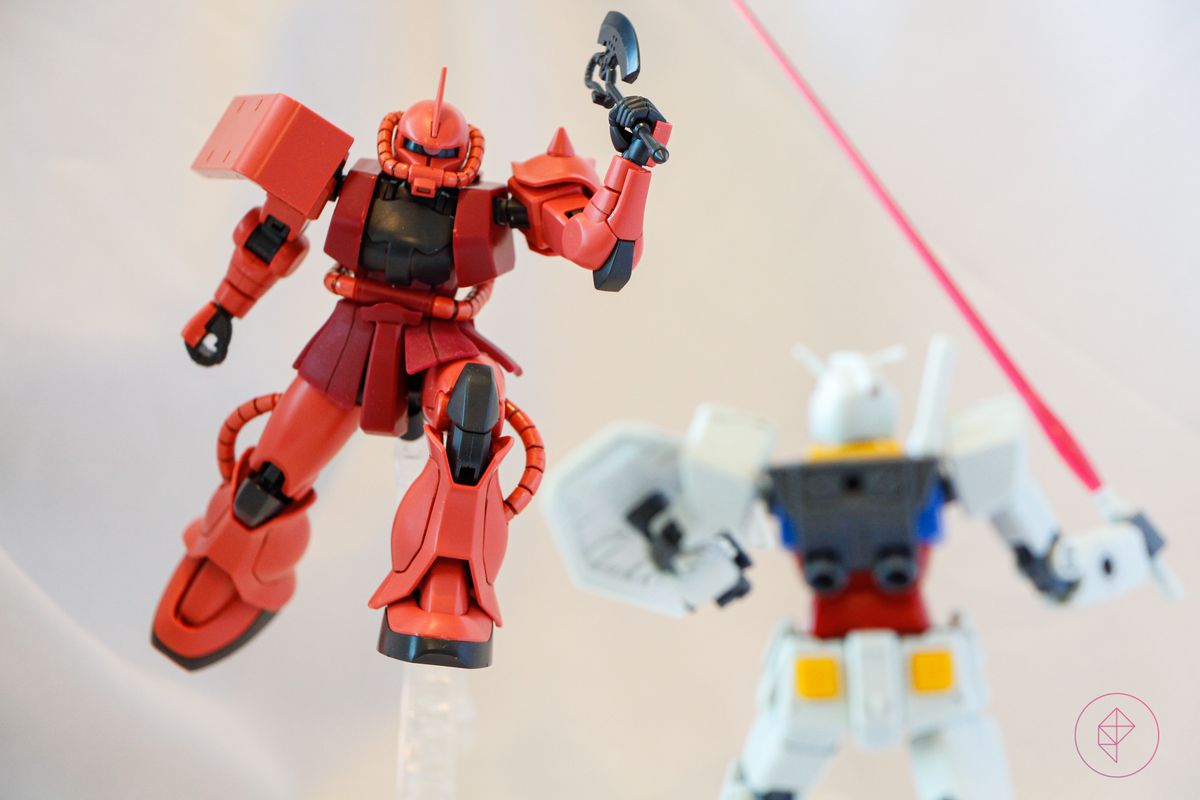 A pink Gunpla flies through the air with the aid of a clear plastic stand, while another gunpla faces away from the camera.