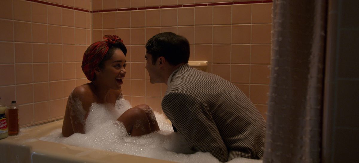 Darren Criss, fully dressed in a suit, crouches in Laura Harrier’s bubble bath with her as she responds eagerly to something he’s saying.