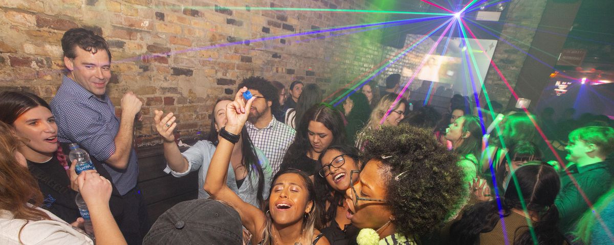 A diverse group of 20-somethings dance and sing inside a club.