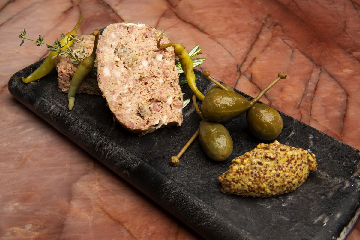 A black slate plate holds a hunk of confit duck rillette beside pickled caper berries.