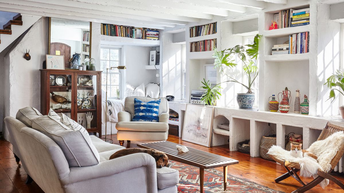 A living room with a grey sofa, coffee table, white bookshelves with books, and multiple windows letting in natural light. There is a patterned area rug on the floor and a white armchair.