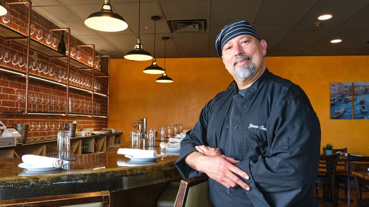 A chef in a black coat poses in front of a bar.
