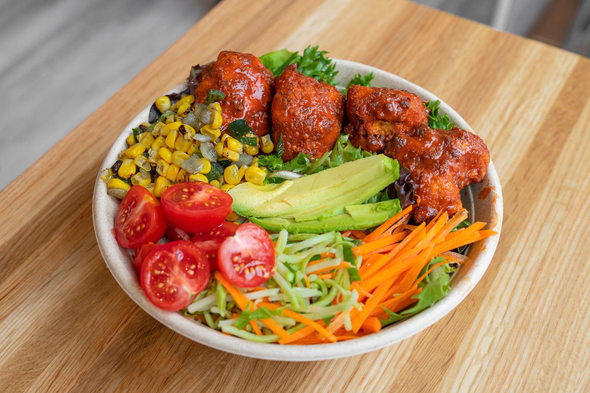 A white bowl filled with Buffalo cauliflower, greens, corn, avocado, and carrots.