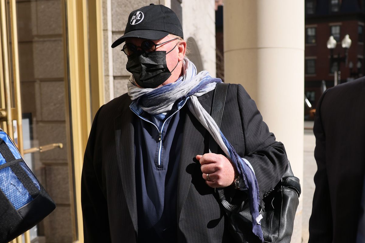 A man in a dark blazer, glasses, baseball cap, face mask, and blue and white scarf carries a shoulder bag and prepares to enter a building.