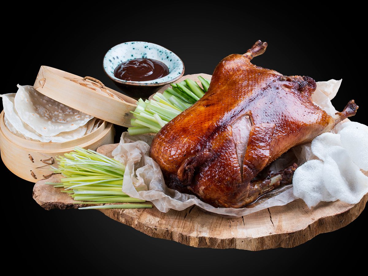 A stock photography of a glossy, dark Peking duck on a wooden platter on a black background, surrounded by accoutrements like Hoisin sauce and a wooden steamer basket of pancakes.