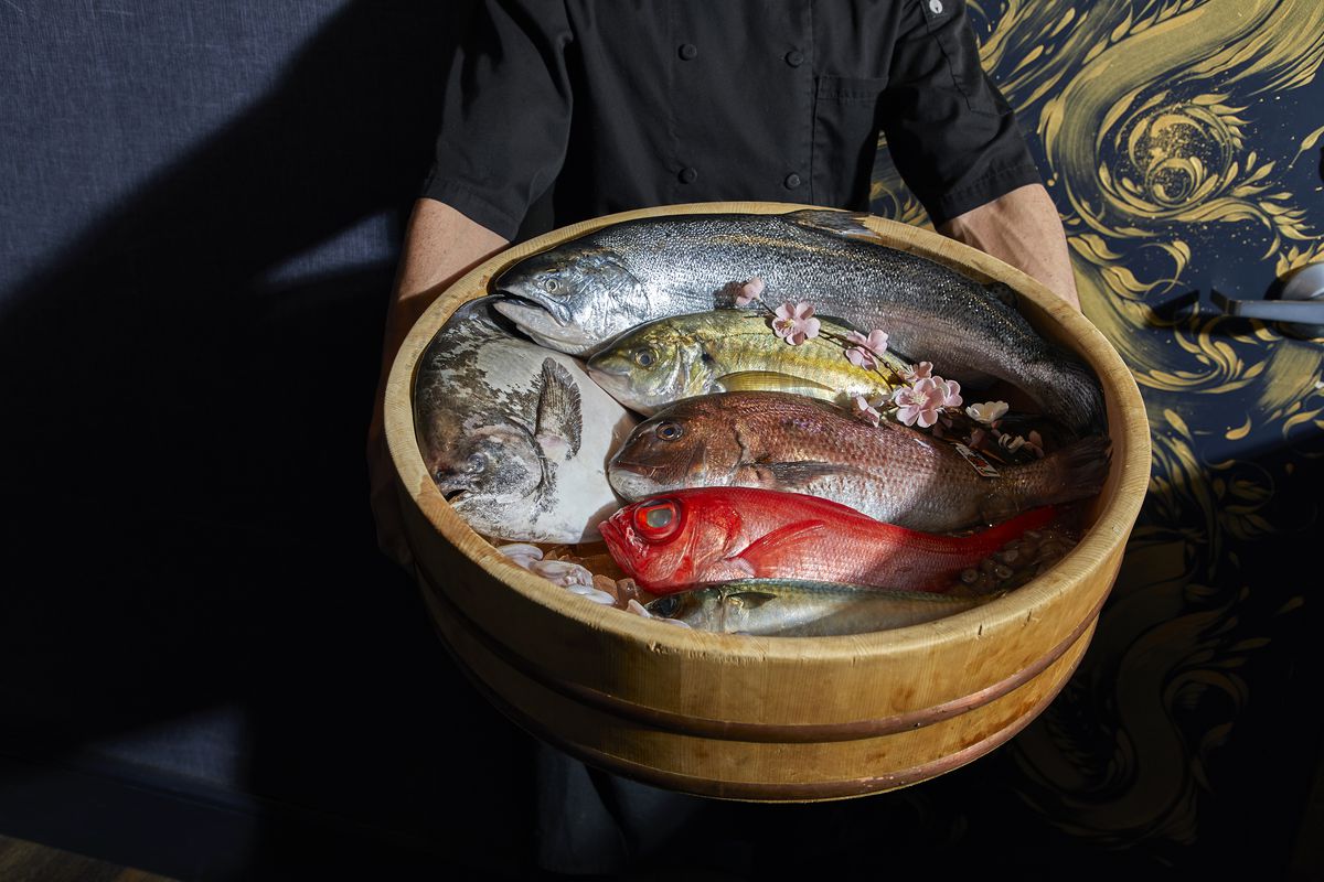 A selection of fresh seafood and fish in a wood basket being held by a person in a black shirt. 