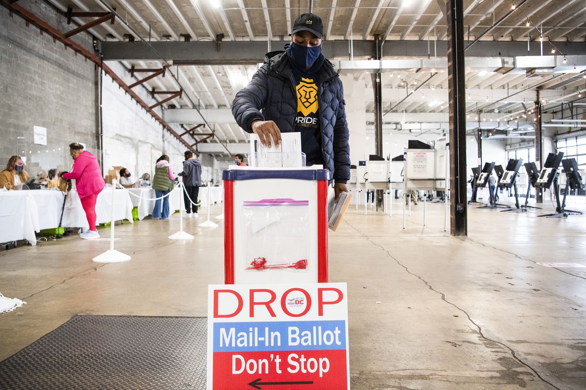 A Black man in a black puffer coat, navy face mask, and black baseball cap drops his ballot into a red and white box. A sign in front of the box reads “DC DROP Mail-In Ballot Don’t Stop.” He is in what appears to be an empty warehouse filled with socially distanced voting booths.