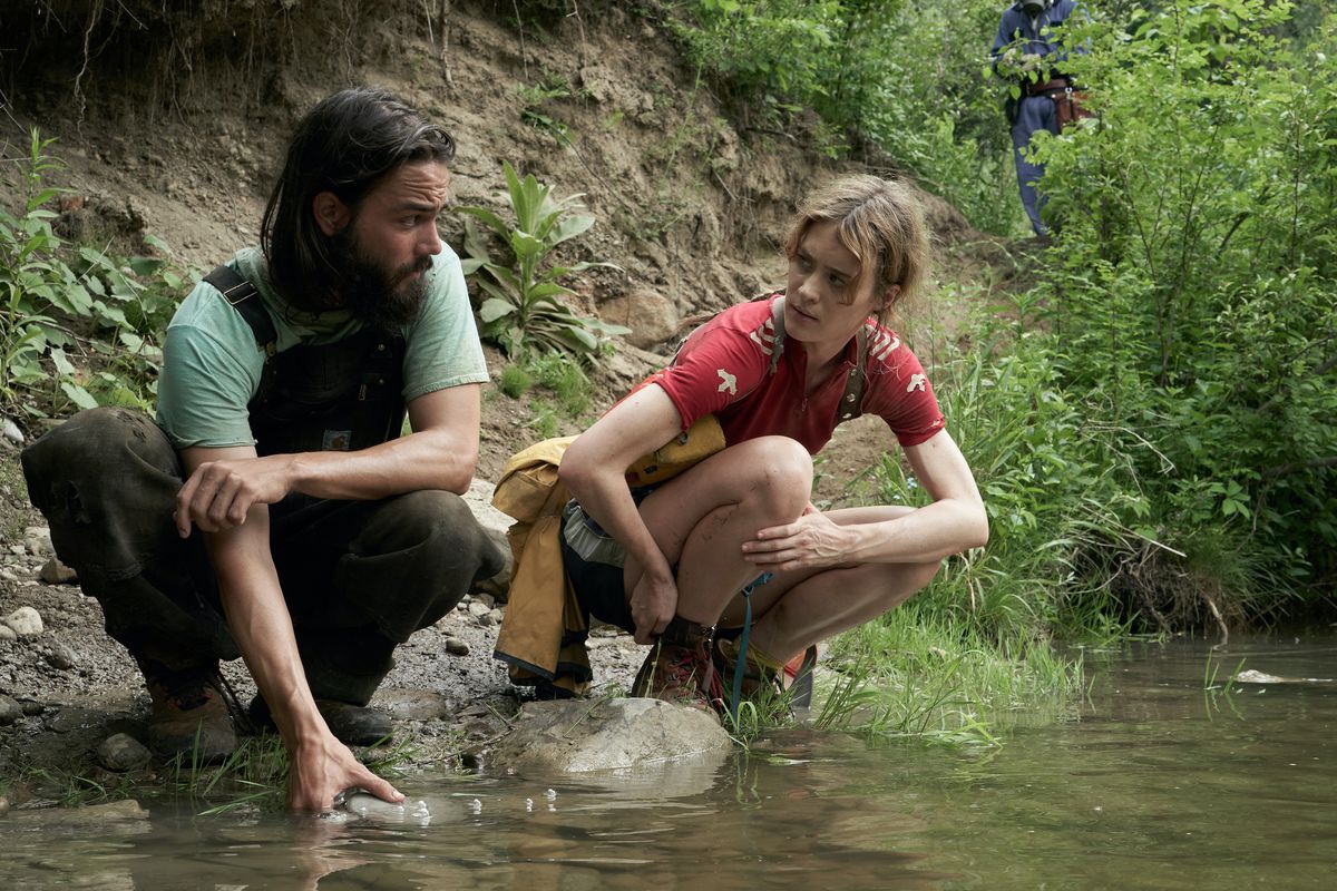 The Prophet and Kirsten stoop by a stream. The image features lush greens and reds.