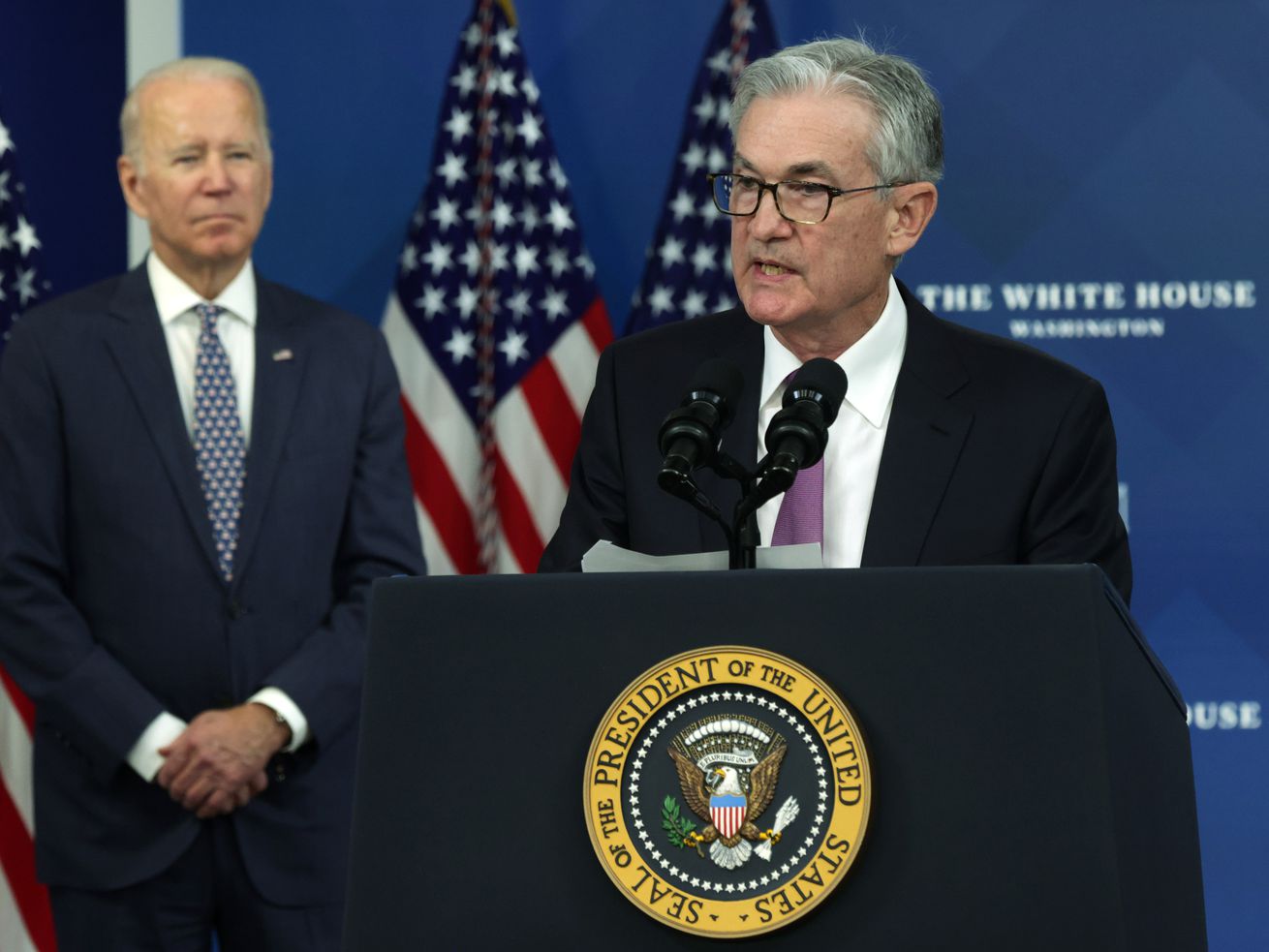 Fed chair Jerome Powell speaks from a lectern as President Joe Biden stands behind him and looks on.