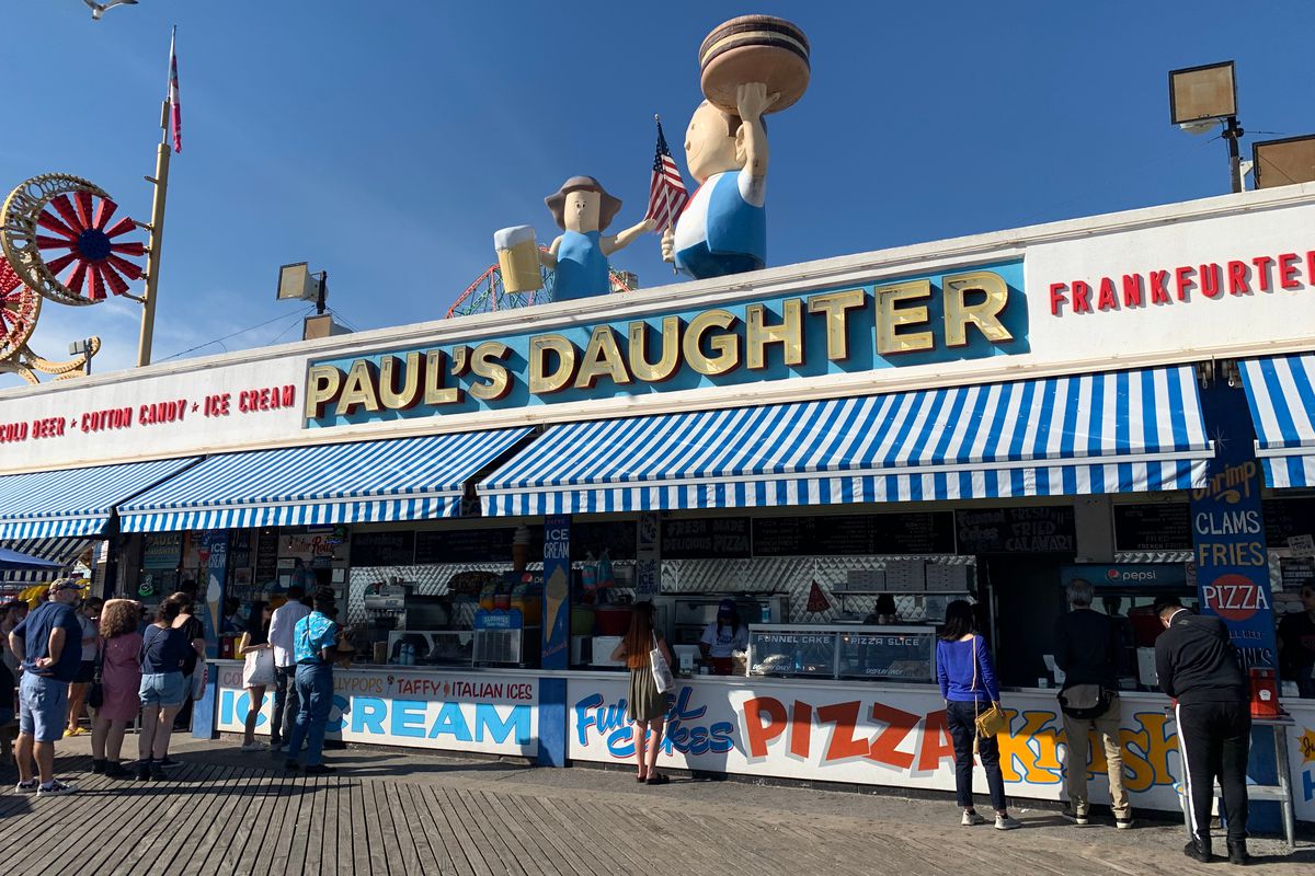 A business next to an amusement park on a boardwalk. The shop has a blue and white striped awning, and a sign that says “Paul’s Daughter.” 