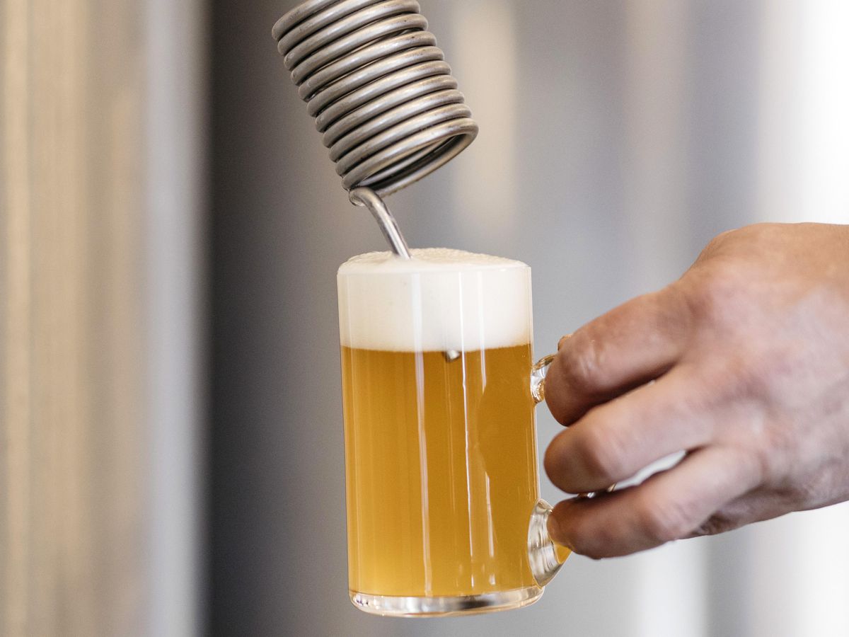 A hand holds a full mug of beer with a thick head of foam beneath a metal tap with a thick spiral just about the mouth