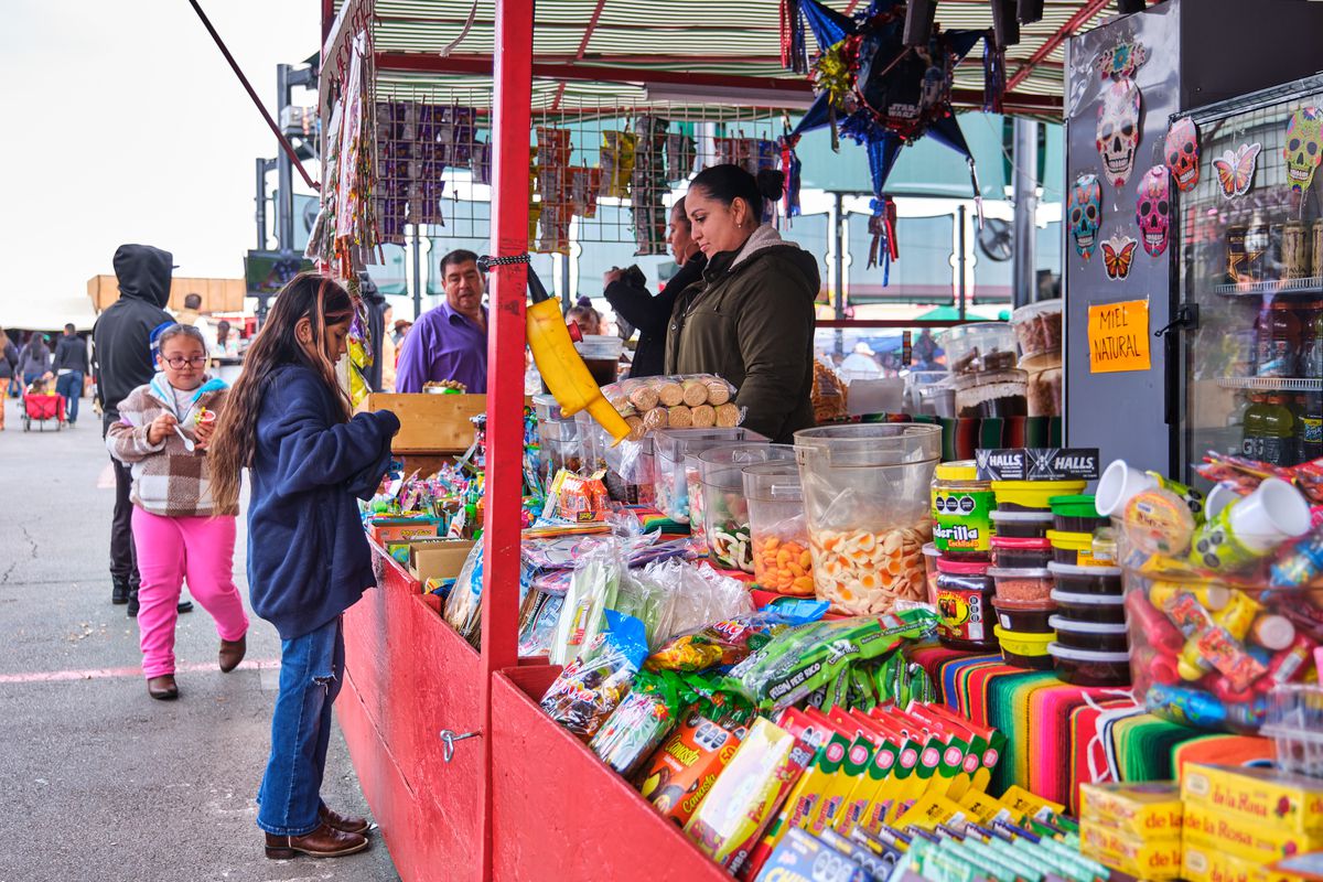 A child, standing in front of a long table covered in gummies, crystallized fruits, caramels, and more, buys candy from an outdoor market.