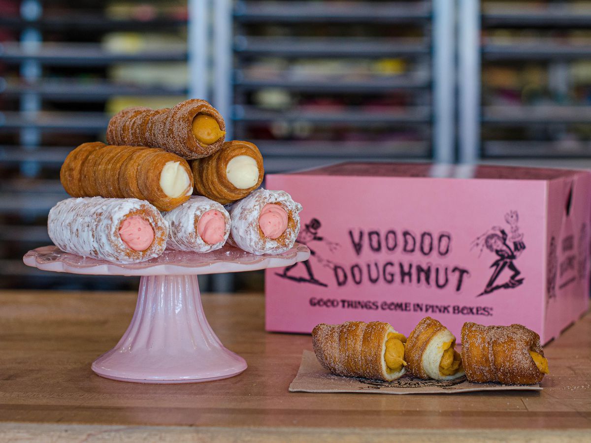 A photo of six tube doughnuts called Cannolos filled with either yellow, brown or pink creme cheese filling and arranged in a stack on a raised plate next to a pink Voodoo Doughnut box