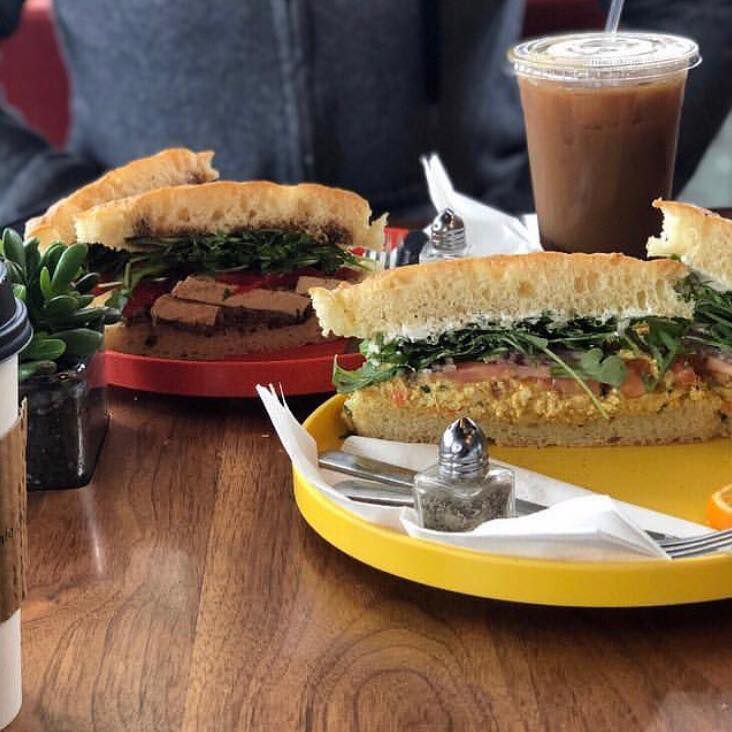 Two sandwiches on colorful plates, sliced in the middle and showing their fillings, face the viewer. An iced coffee drink is in the background.