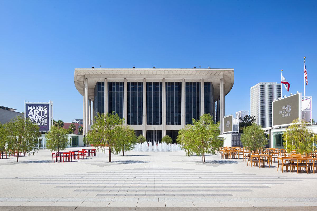 A flat open outdoor plaza with seating at scattered tables. Eight young trees are also on the plaza. In the background is the Dorothy Chandler Pavilion, a modern building with a concrete skeleton and a flat white roof.