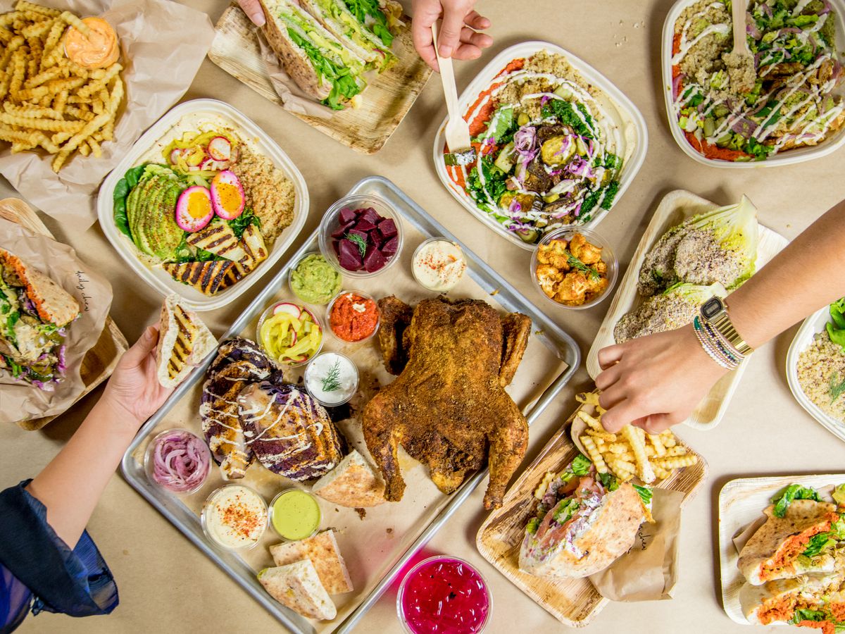 Hands reach out to sample a takeout spread of hummus, pita, a whole seasoned rotisserie chicken, chicken salad, pita with za’taar, and fries.