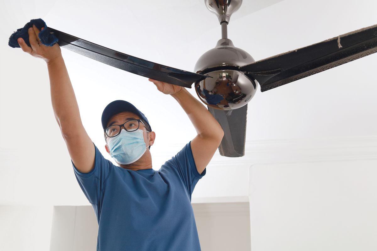 Man wearing a mask cleaning the dust off of a ceiling fan. 