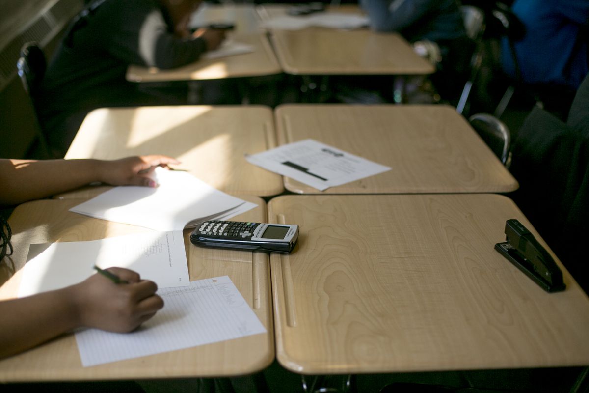 In a close-up of a student’s hands in math class, worksheets and a calculator are spread across the desk.