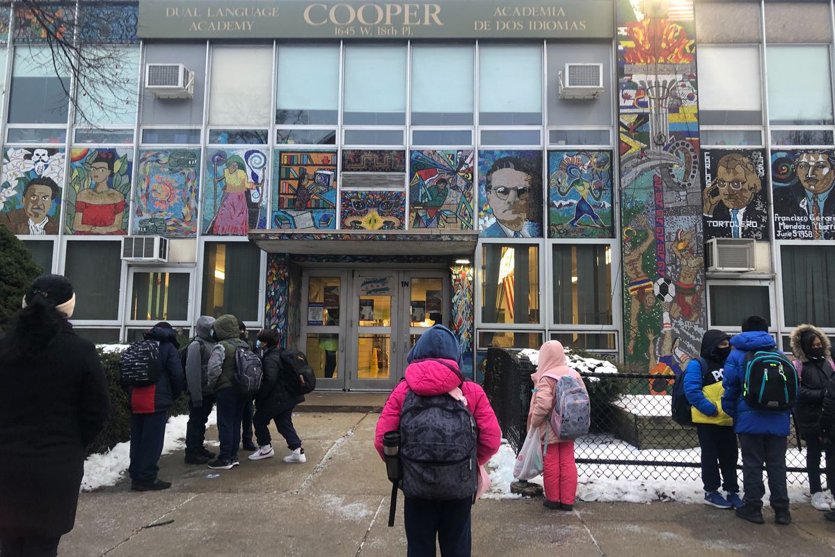 Students, wearing masks and carrying backpacks, accompanied by parents, line up along outside Cooper Elementary in Pilsen after a week of canceled classes.