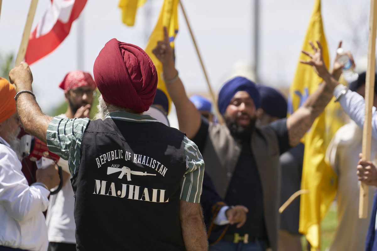 Sikhs holding flags at an outdoor protest, and one person with their back to the camera has a vest with “Republic of Khalistan” on it.