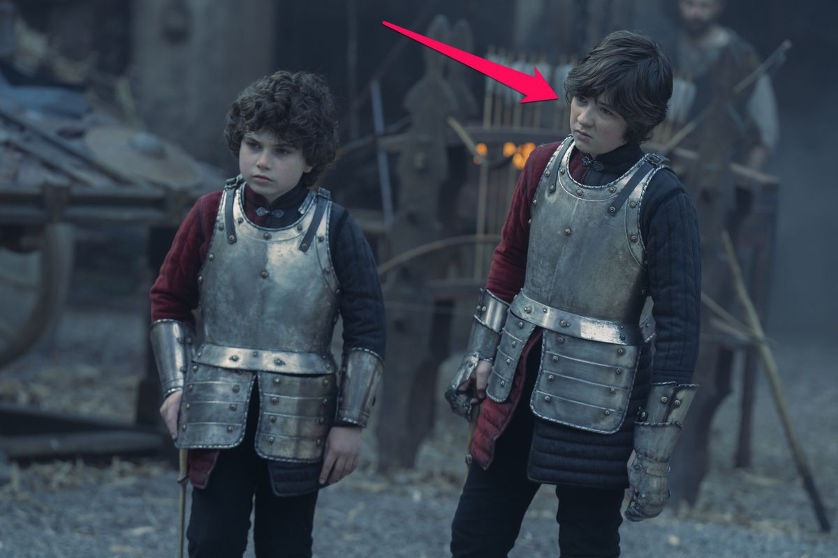 Leo Hart as Jacaerys Valeryon from House of the Dragon. A young boy with longer brown hair stands next to his brother, who has curly brown hair. Both wear chest armor. An arrow points to Jacaerys, who has longer hair.