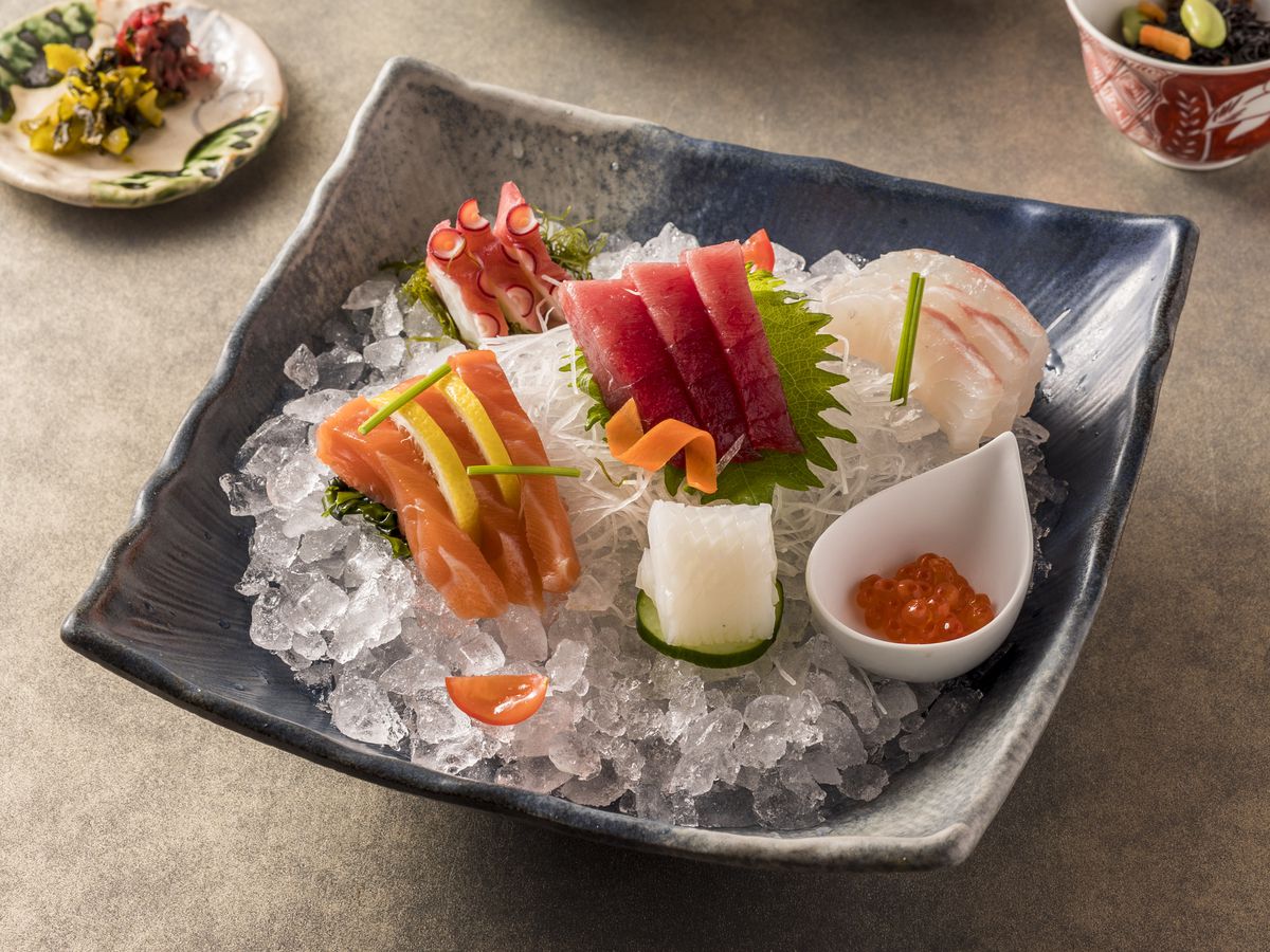 A square slate bowl of sliced sashimi on ice, beside accompaniments like rice, pickles, and tea
