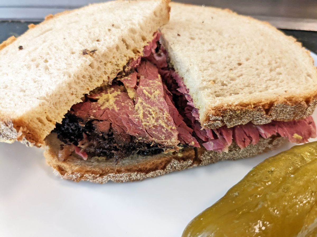 A sandwich on rye cut in half and filled with red meat, with a pickle in the foreground.