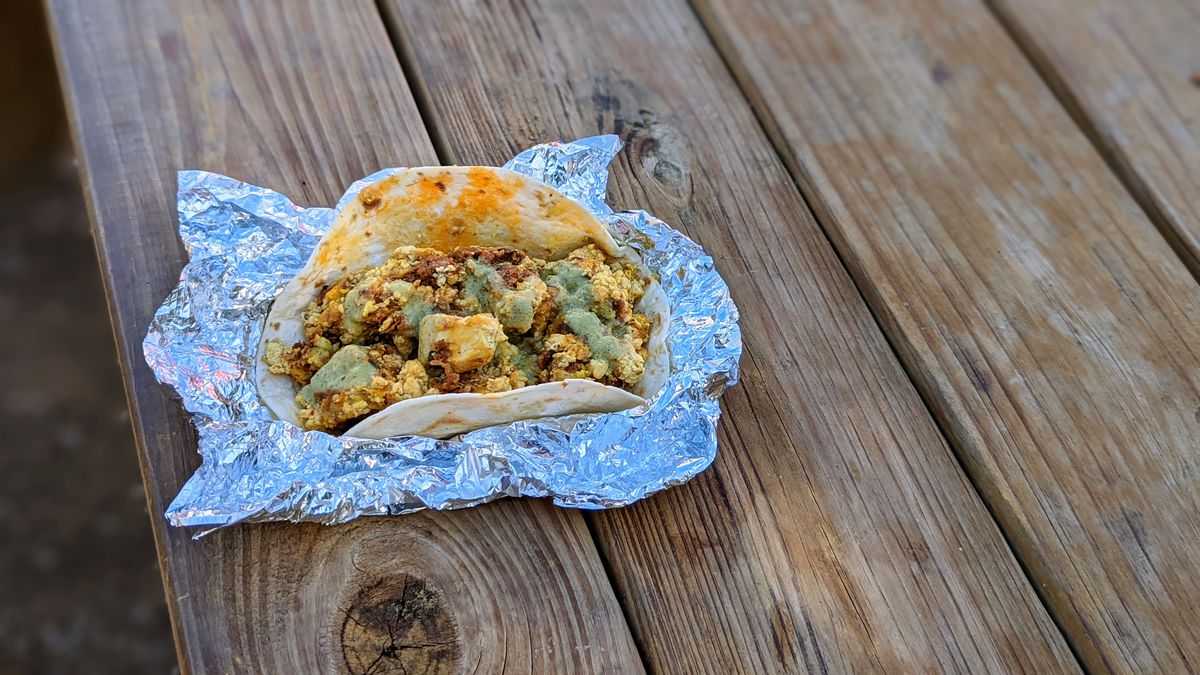 A taco with eggs, vegetables, and a brown sauce in an opened foil wrapper on a wooden table.